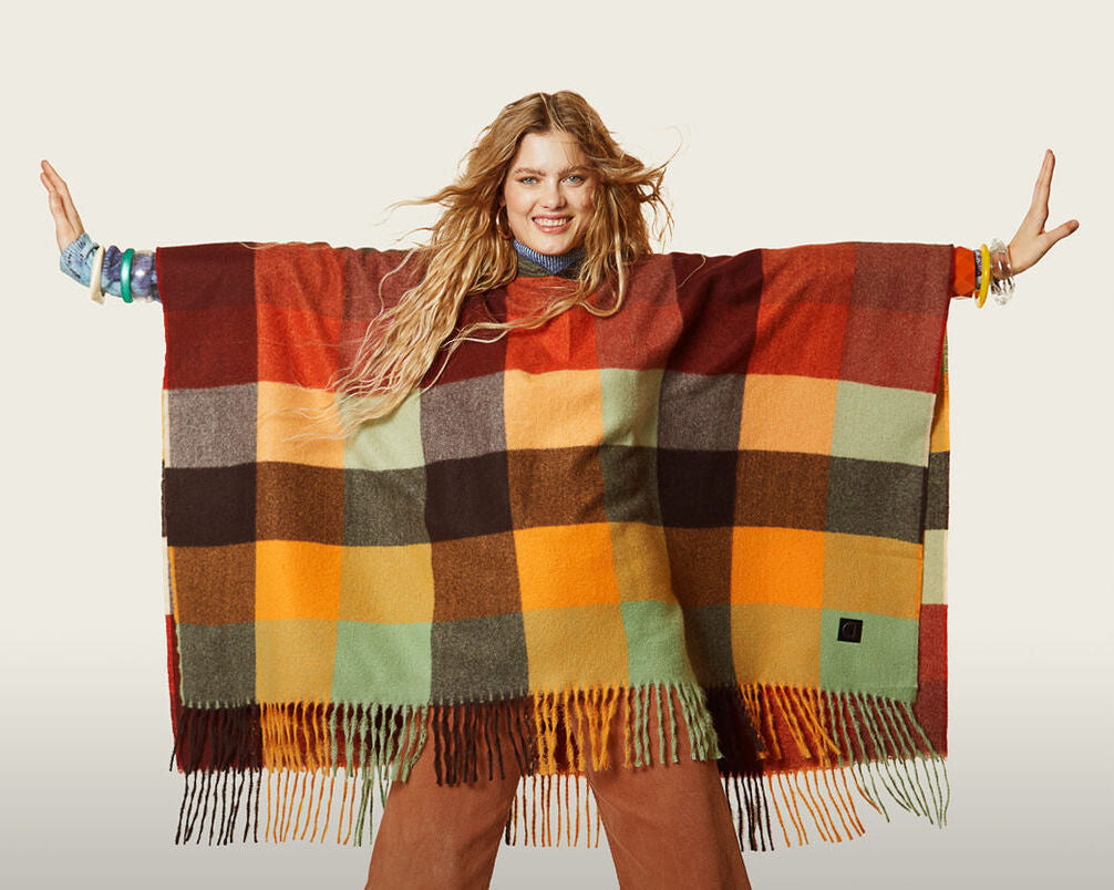 Front top half view of a woman wearing the desigual plaid poncho with brown pants. This poncho is brown, red, orange, and green plaid. It has a hood and a fringed hem.