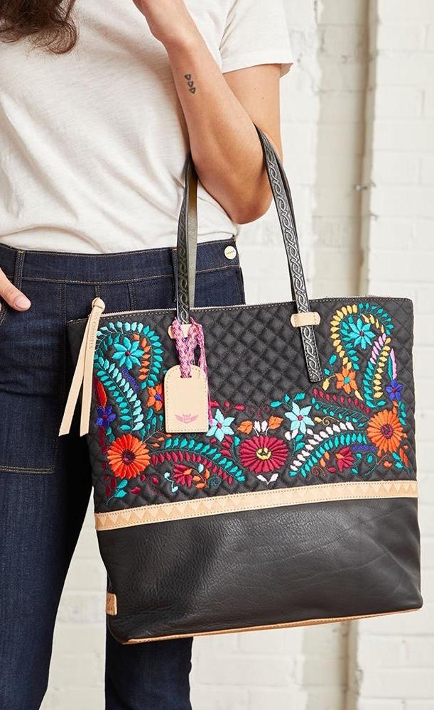 Front view of a woman carrying the consuela venice market tote. This tote is black with a quilted top half and light tan trim. The straps are thin. On the front of the tote is blue pink and orange floral embroidery.
