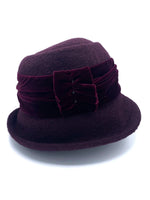 Load image into Gallery viewer, left side view of the lillie &amp; cohoe aubergine wool jeanette hat. This hat is wine/purple colored and has a rounded brim and a velvet band that wraps around the crown to make a bow on the side.

