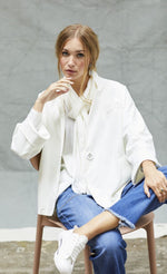 Load image into Gallery viewer, Front view of a woman sitting on a chair wearing blue jeans and the white indies voyageur jacket. This jacket is open and layered over a white top with a with scarf. The jacket has a single front button.
