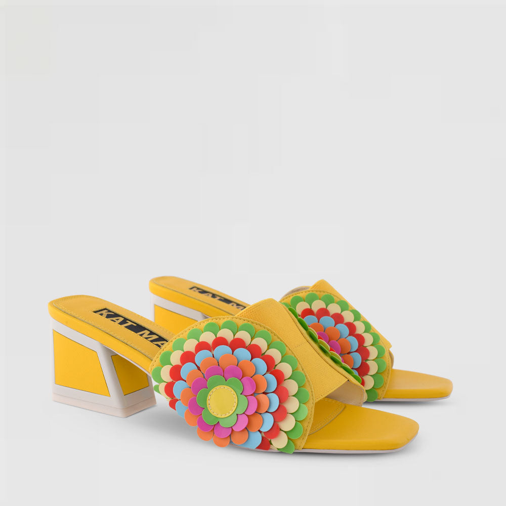 Front outer side view of the kat maconie vira kicker heel sandals. These mule sandals are mineral yellow with multi colored flowers on the sides. These sandals also have a block heel and an open toe front.