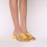 Load image into Gallery viewer, Front outer side view of a woman wearing a pair of the kat maconie vira kicker heel sandals. These mule sandals are mineral yellow with multi colored flowers on the sides. These sandals also have a block heel and an open toe front.
