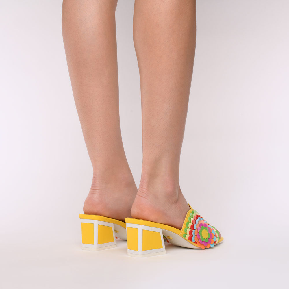 Back view of a woman wearing a pair of the kat maconie vira kicker heel sandals. These mule sandals are mineral yellow with multi colored flowers on the sides. These sandals also have a block heel and an open toe front.