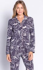 Load image into Gallery viewer, Front top half view of a woman wearing the pj salvage love lines set. This pj set is charcoal colored with white flowers drawn all over it and light pink detailing. The top has a button down front and long sleeves. The bottom is relaxed.

