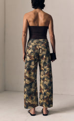 Load image into Gallery viewer, A model shown from the back wearing wide-leg cropped pants with an abstract watercolor-style pattern in olive green, brown, beige, and black, paired with a black strapless top and black sandals.
