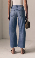 Load image into Gallery viewer, Person wearing blue jeans and a white top, holding a brown bag on a light background
