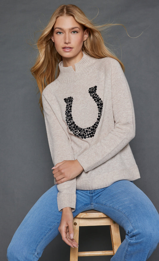 Front top half view of a woman wearing the lisa todd lucky U sweater in oat with a stone embellished horse shoe.