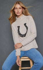 Load image into Gallery viewer, Front top half view of a woman wearing the lisa todd lucky U sweater in oat with a stone embellished horse shoe.
