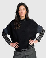 Load image into Gallery viewer, A woman modeling a black knit poncho-style sweater with leopard-print sleeves and a matching leopard-print hem, paired with gray trousers, against a light gray studio background.
