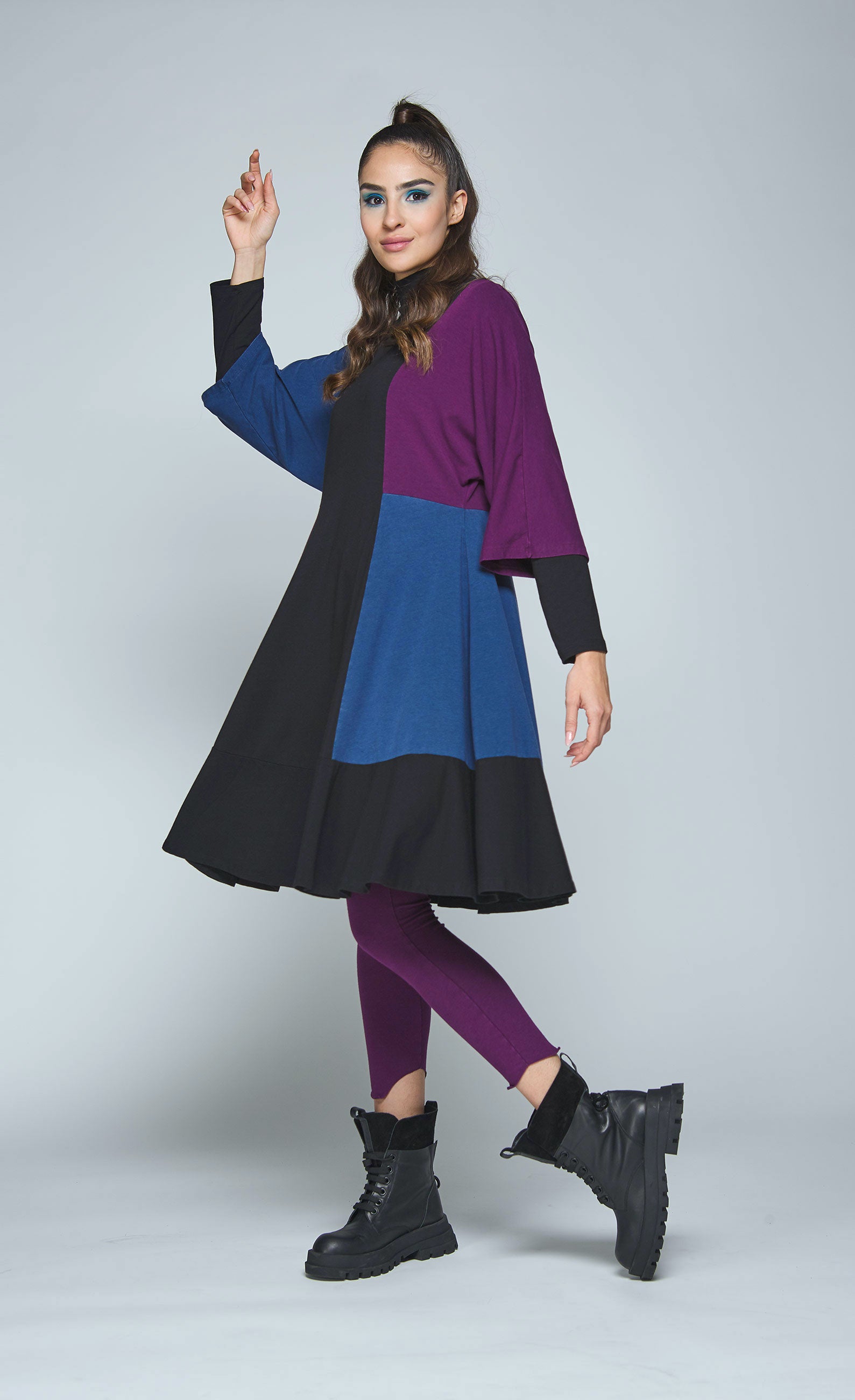 Left side full body view of a woman wearing the luukaa color block tunic/dress. This knee-length dress is black with a blue and purple color blocking on the sides and sleeves. The dress also has elbow-length sleeves and a funnel neck.