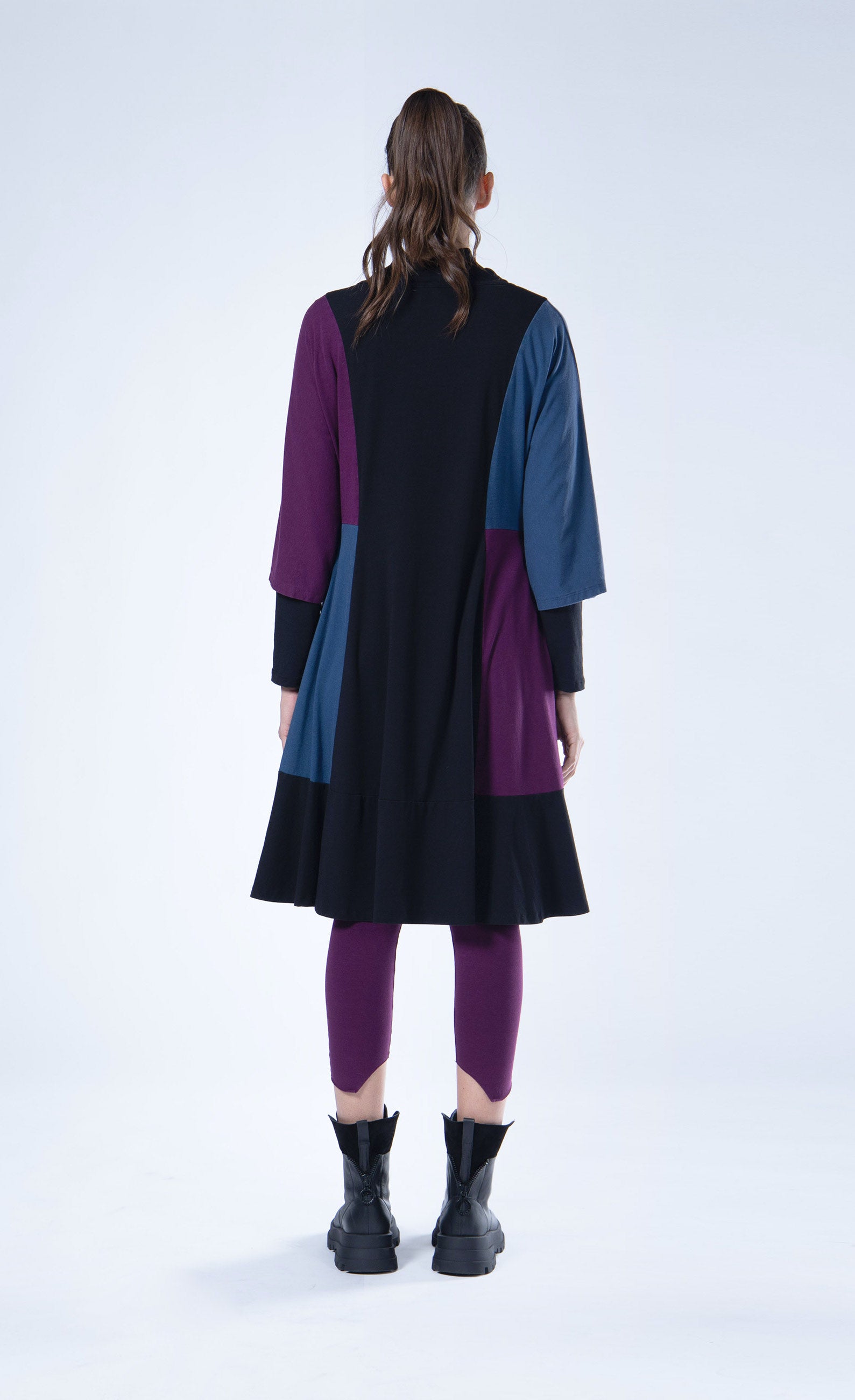 Back full body view of a woman wearing the luukaa color block tunic/dress. This knee-length dress is black with a blue and purple color blocking on the sides and sleeves. The dress also has elbow-length sleeves and a funnel neck.