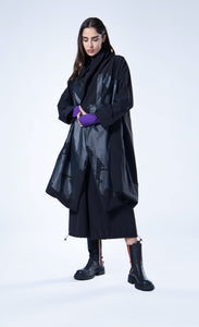 Front full body view of a woman wearing the luukaa draped open black coat. This coat is black with a black contrast, glossy abstract print on the front. The coat has long sleeves, a billowy fit, and a draped open front with a hem that sits at the knees.