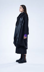 Load image into Gallery viewer, Left side full body view of a woman wearing the luukaa draped open black coat. This coat is black with a black contrast, glossy abstract print on the front. The coat has long sleeves, a billowy fit, and a draped open front with a hem that sits at the knees.
