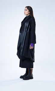 Left side full body view of a woman wearing the luukaa draped open black coat. This coat is black with a black contrast, glossy abstract print on the front. The coat has long sleeves, a billowy fit, and a draped open front with a hem that sits at the knees.