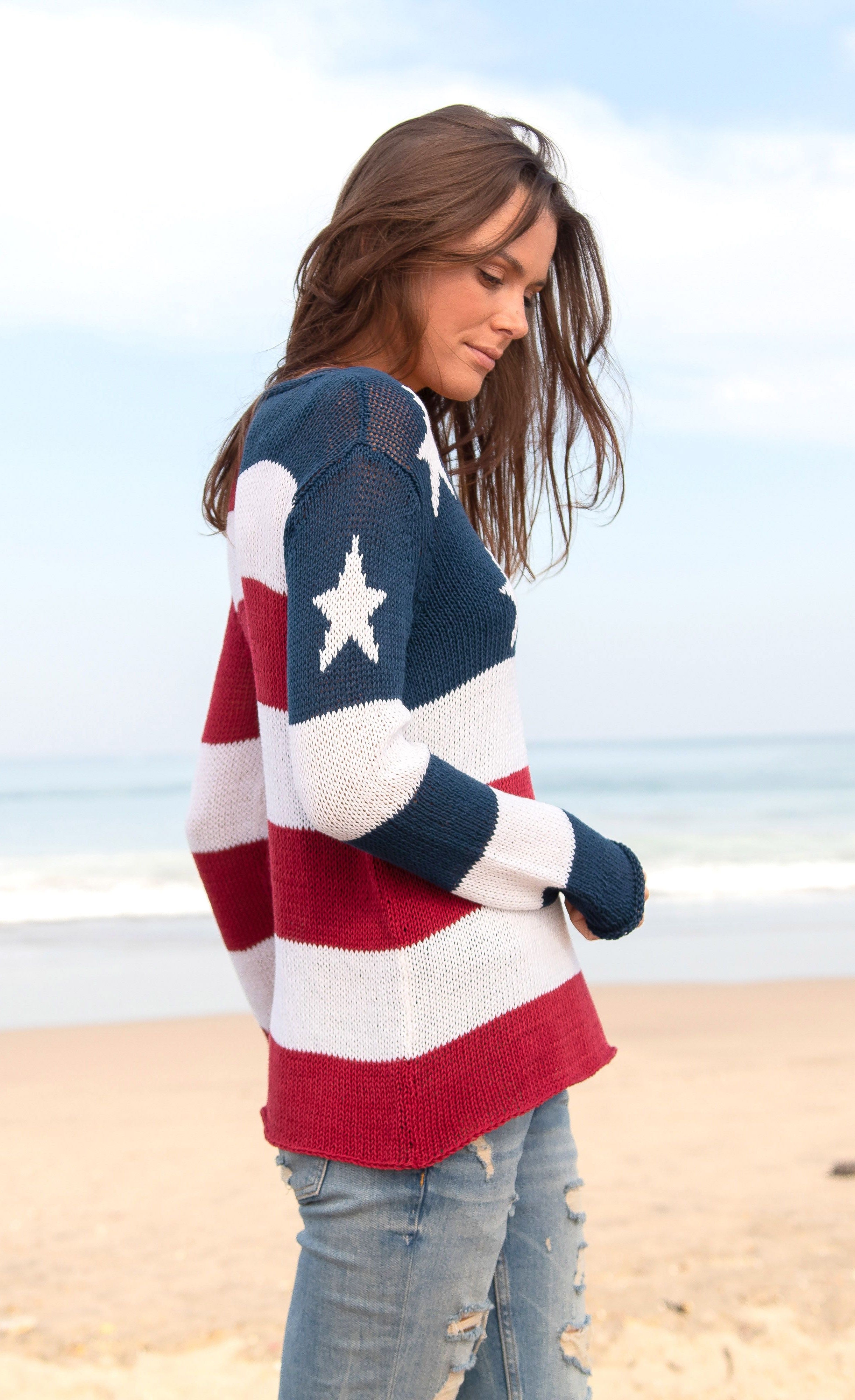 Right side top half view of a woman standing on the beach wearing the wooden ships flag v cotton sweater. This sweater has the flag on it with a v-neck and long sleeves.