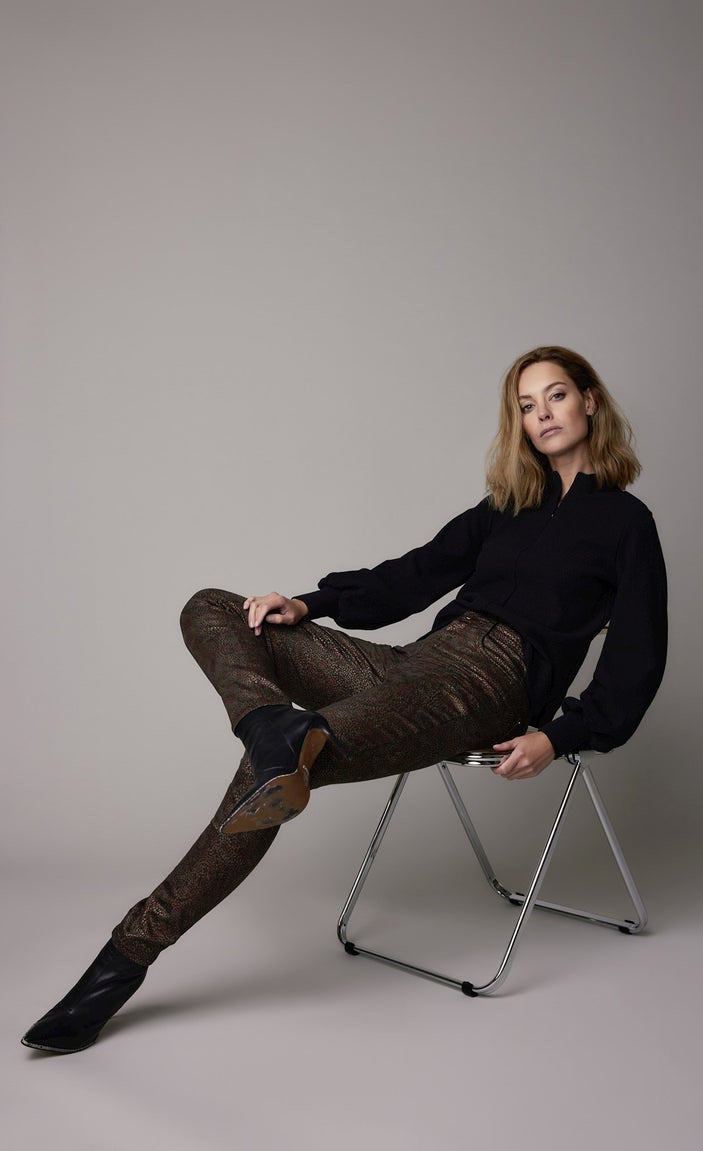 Front full body view of a woman sitting and wearing a black top and the summum Skinny trousers with foil print. This pant is caramel bronze colored with a small metallic gold and brown leaf print all over it.