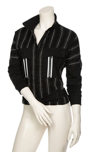 Front, top half view of a mannequin wearing the Beate Heymann Stripped Jacket. This jacket is black with white stripes. The sleeves are solid black and the front has two patch, solid black pockets with a single white stripe on them. The jacket has a zip up front and belt near the bottom.