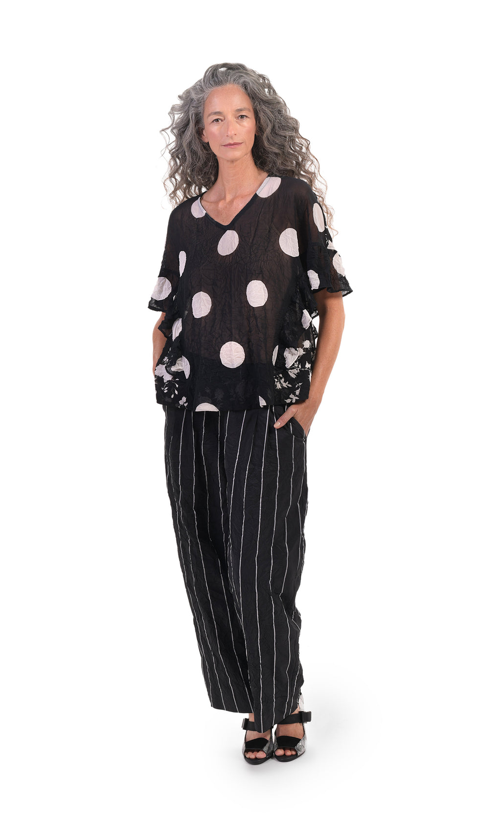 Front full body view of a woman wearing striped pants and the alembika dot floral crinkled top. This top is black with white dots on the front and white floral print on the back and sides. The shirt has a v-neck and short, elbow length sleeves with ruffles that run down the sides. 