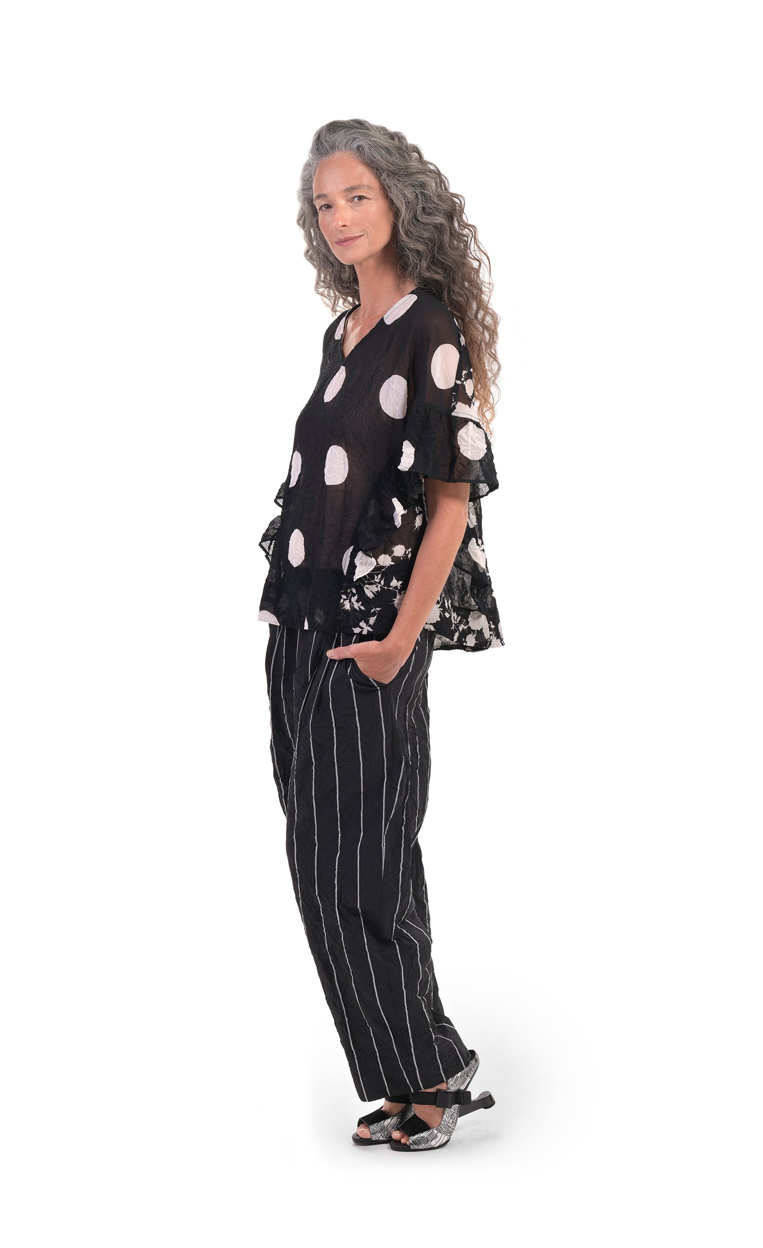Left sided, full body view of a woman wearing striped pants and the alembika dot floral crinkled top. This top is black with white dots on the front and white floral print on the back and sides. The shirt has a v-neck and short, elbow length sleeves with ruffles that run down the sides. 