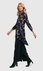 Load image into Gallery viewer, Full body left side view of a woman wearing the alembika amethyst crinkle tunic blouse. This tunic is tan with black, purple, and blue large animal print on it. The top has a button down front, a collar, crinkle fabric, and long sleeves.
