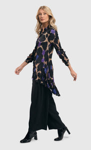 Full body left side view of a woman wearing the alembika amethyst crinkle tunic blouse. This tunic is tan with black, purple, and blue large animal print on it. The top has a button down front, a collar, crinkle fabric, and long sleeves.