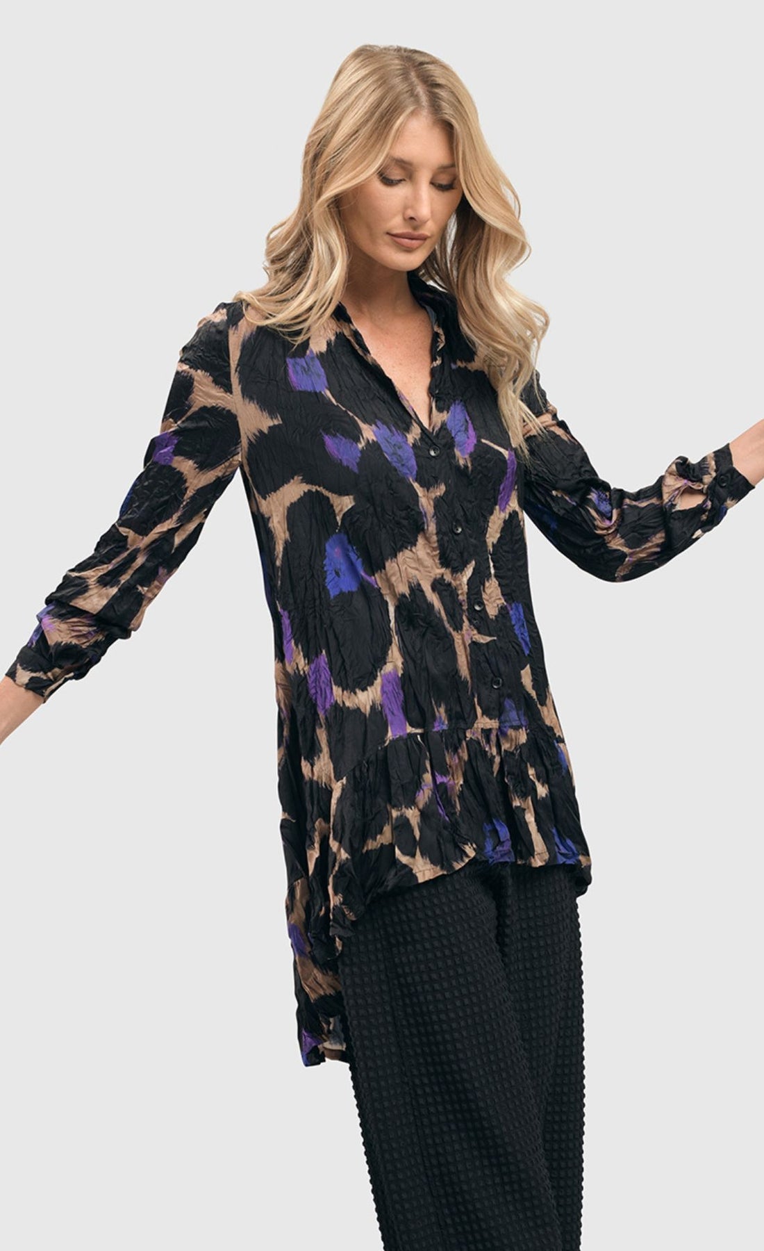 Front top half view of a woman wearing the alembika amethyst crinkle tunic blouse. This tunic is tan with black, purple, and blue large animal print on it. The top has a button down front, a collar, crinkle fabric, and long sleeves.