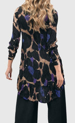 Load image into Gallery viewer, Back top half of a woman wearing the alembika amethyst crinkle tunic blouse. This tunic is tan with black, purple, and blue large animal print on it. The top has a button down front, a collar, crinkle fabric, and long sleeves.
