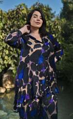 Load image into Gallery viewer, Front view of a woman wearing the alembika amethyst crinkle tunic blouse. This tunic is tan with black, purple, and blue large animal print on it. The top has a button down front, a collar, crinkle fabric, and long sleeves.
