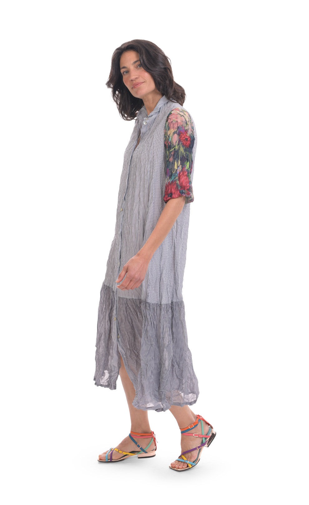 Left side full body view of a woman wearing the alembika flora dress. This dress has navy and white striping, a button down front, and floral 3/4 sleeves. The bottom 1/4 of the dress is more transparent than the top 3/4.