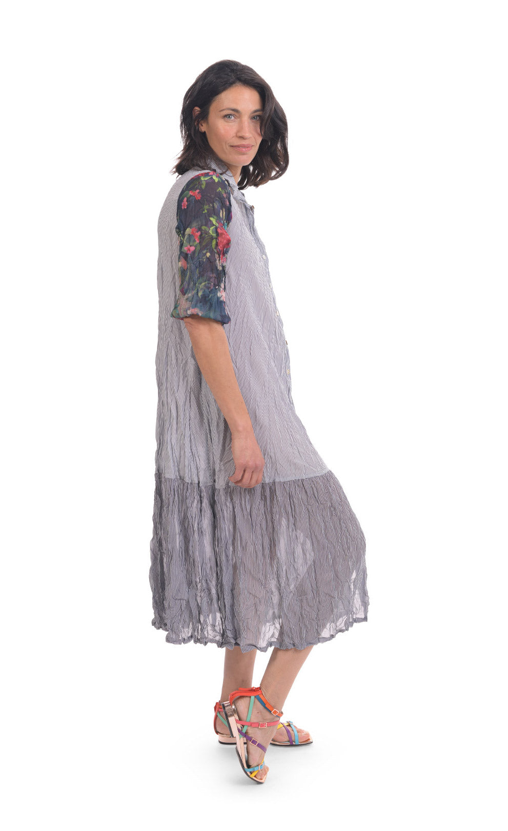 Right side full body view of a woman wearing the alembika flora dress. This dress has navy and white striping, a button down front, and floral 3/4 sleeves. The bottom 1/4 of the dress is more transparent than the top 3/4.