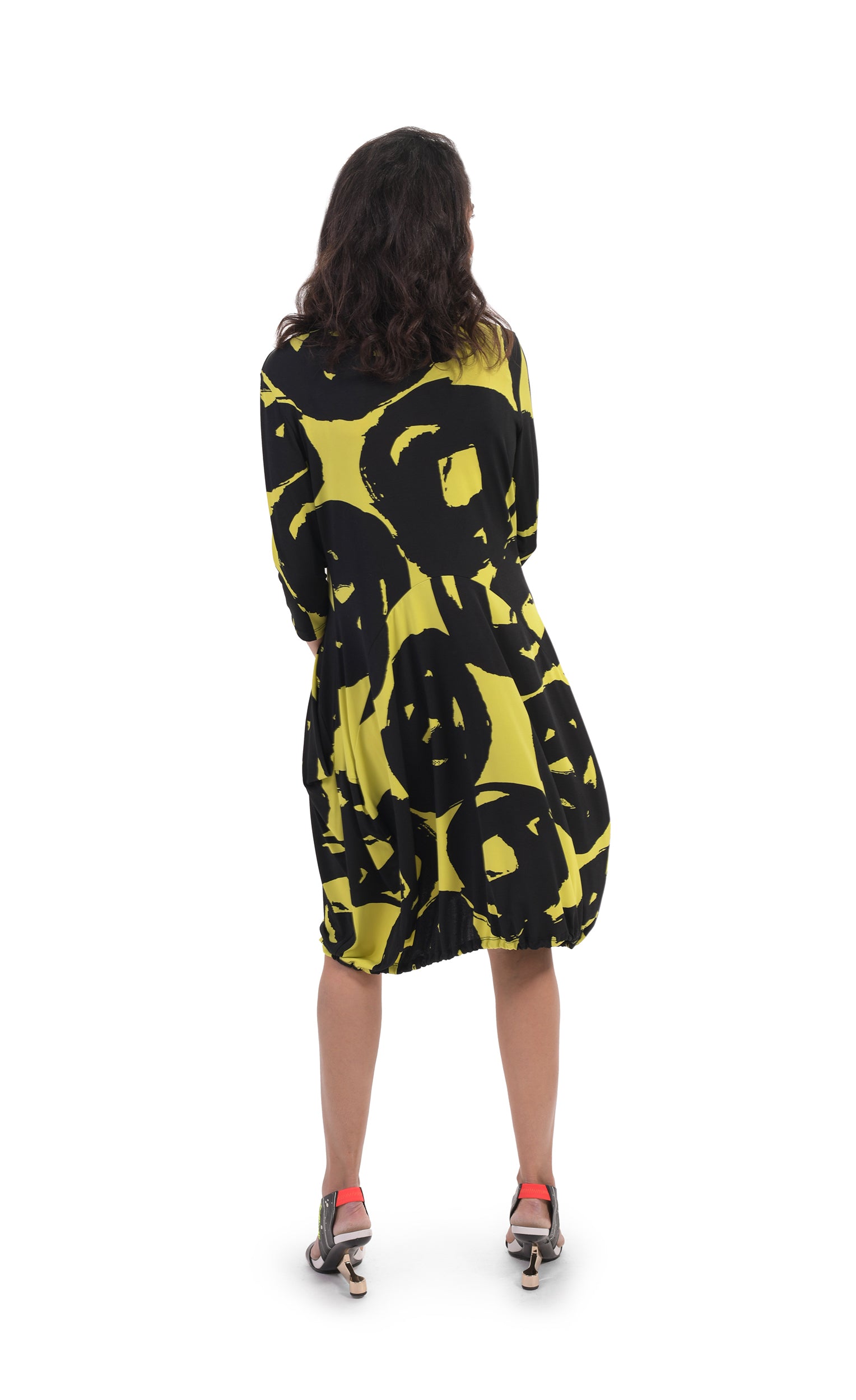 Back full body view of a woman wearing the alembika art print wonderful collar dress. This dress is tapered in at the torso with a poof skirt. It is lime with a black swirl print. 