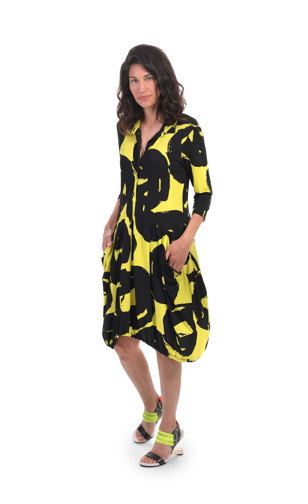 Front full body view of a woman wearing the alembika art print wonderful collar dress. This dress is tapered in at the torso with a poof skirt. It is lime with a black swirl print. The front has two pockets on the skirt, a half button front, and 3/4 length sleeves.