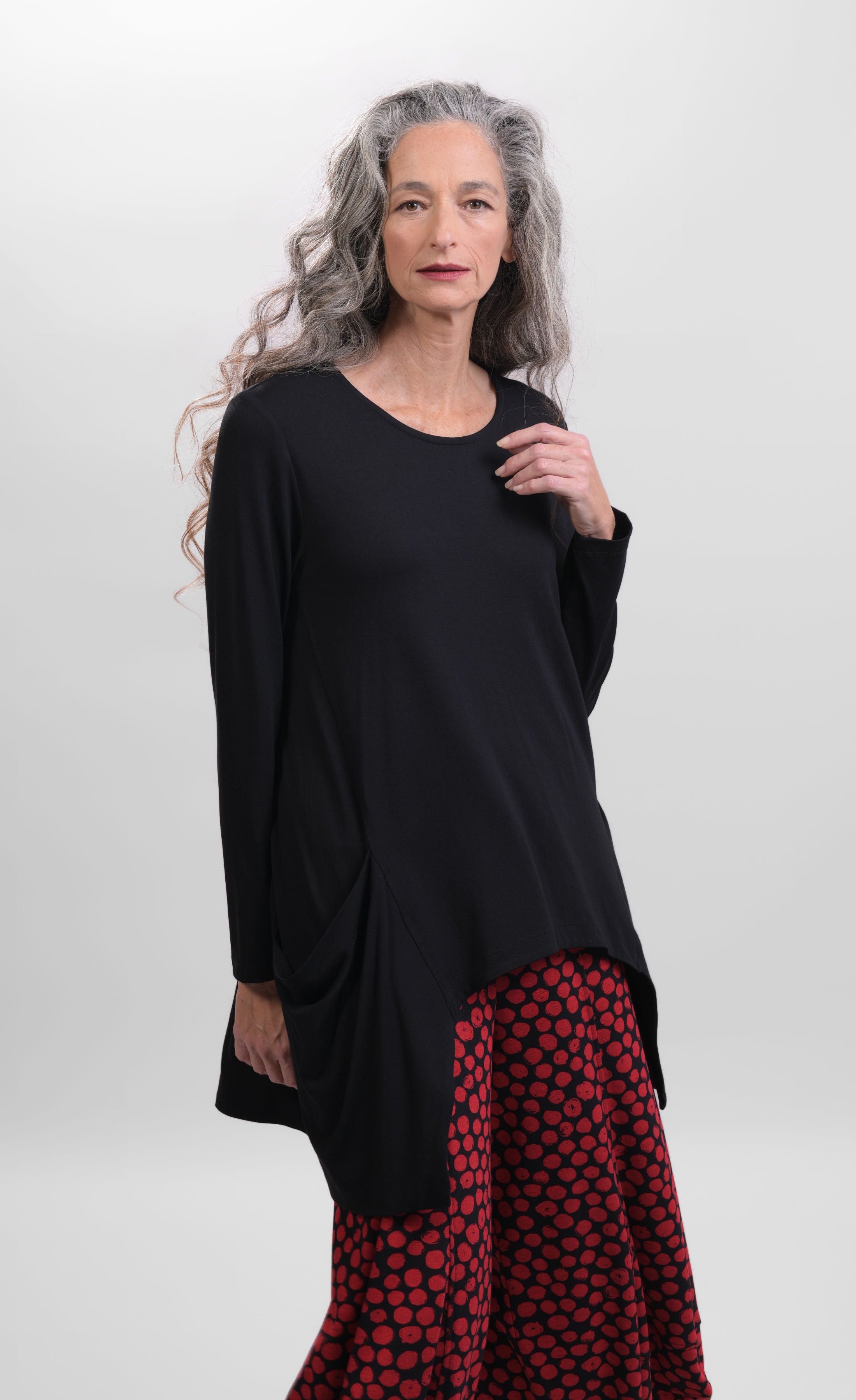 Front top half view of a woman wearing the Alembika Pocket Top in black. This top has long sleeves and two front draped pockets that are longer than the front hem. 