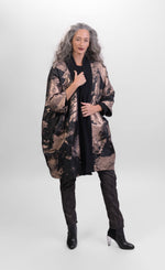 Load image into Gallery viewer, Front full body view of a woman wearing pants and the Alembika Bronze Jacket. This jacket is black with a metallic bronze print on it. The jacket goes down to the knees and has 3/4 length sleeves, two front pockets, and a single button front.
