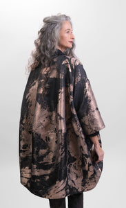 Back top half view of a woman wearing pants and the Alembika Bronze Jacket. This jacket is black with a metallic bronze print on it. The jacket goes down to the knees and has 3/4 length sleeves and an oversized fit.