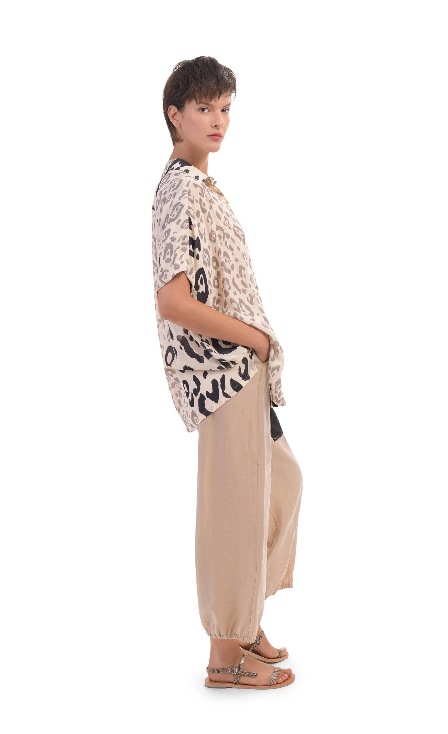 Right side full body view of a woman wearing the alembika Cheetah Print Button Down Top. This top has different panels of black and taupe cheetah print on a creme background. The top has short dolman sleeves. On the bottom the woman is wearing cream pants.