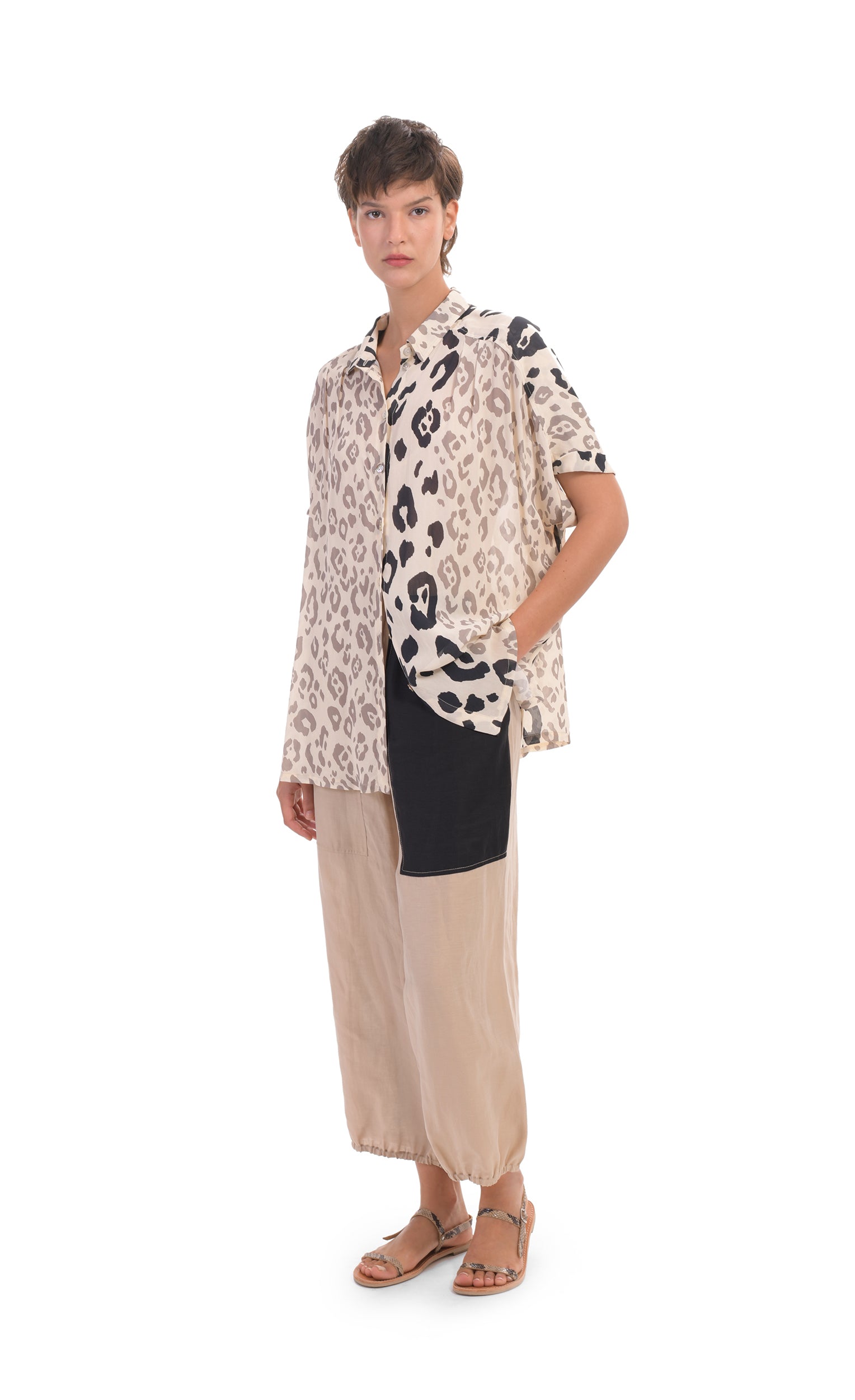Front full body view of a woman wearing the alembika Cheetah Print Button Down Top. This top has different panels of black and taupe cheetah print on a creme background. The top has a button down front, short dolman sleeves, and a back pleat. on the bottom the woman is wearing cream pants with a black patch pocket.