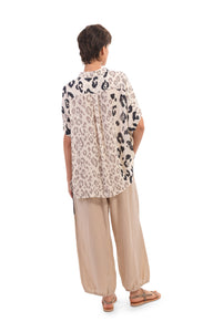 Back full body view of a woman wearing the alembika Cheetah Print Button Down Top. This top has different panels of black and taupe cheetah print on a creme background. The top has short dolman sleeves and a back pleat. On the bottom the woman is wearing cream pants.