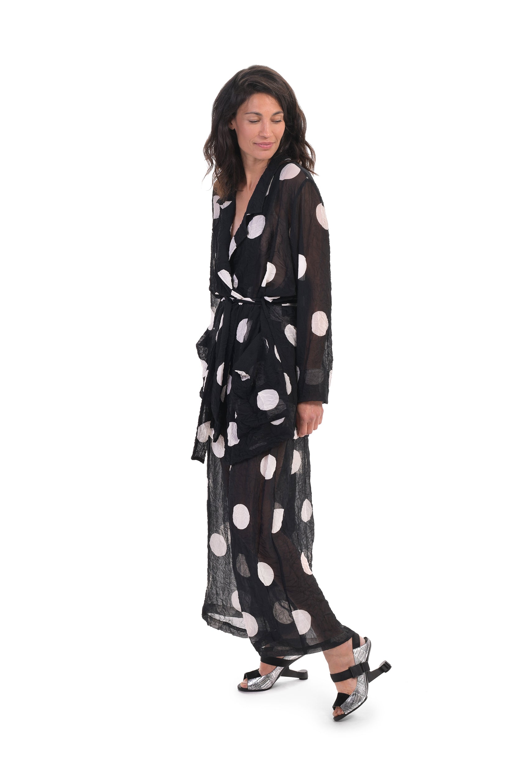 Left side full body view of a woman wearing the alembika chiffon dot palazzo pant with a matching chiffon shirt. The pant and shirt is black with white dots. The fabric appears sheer. The pants are wide legged and end right above the ankles.