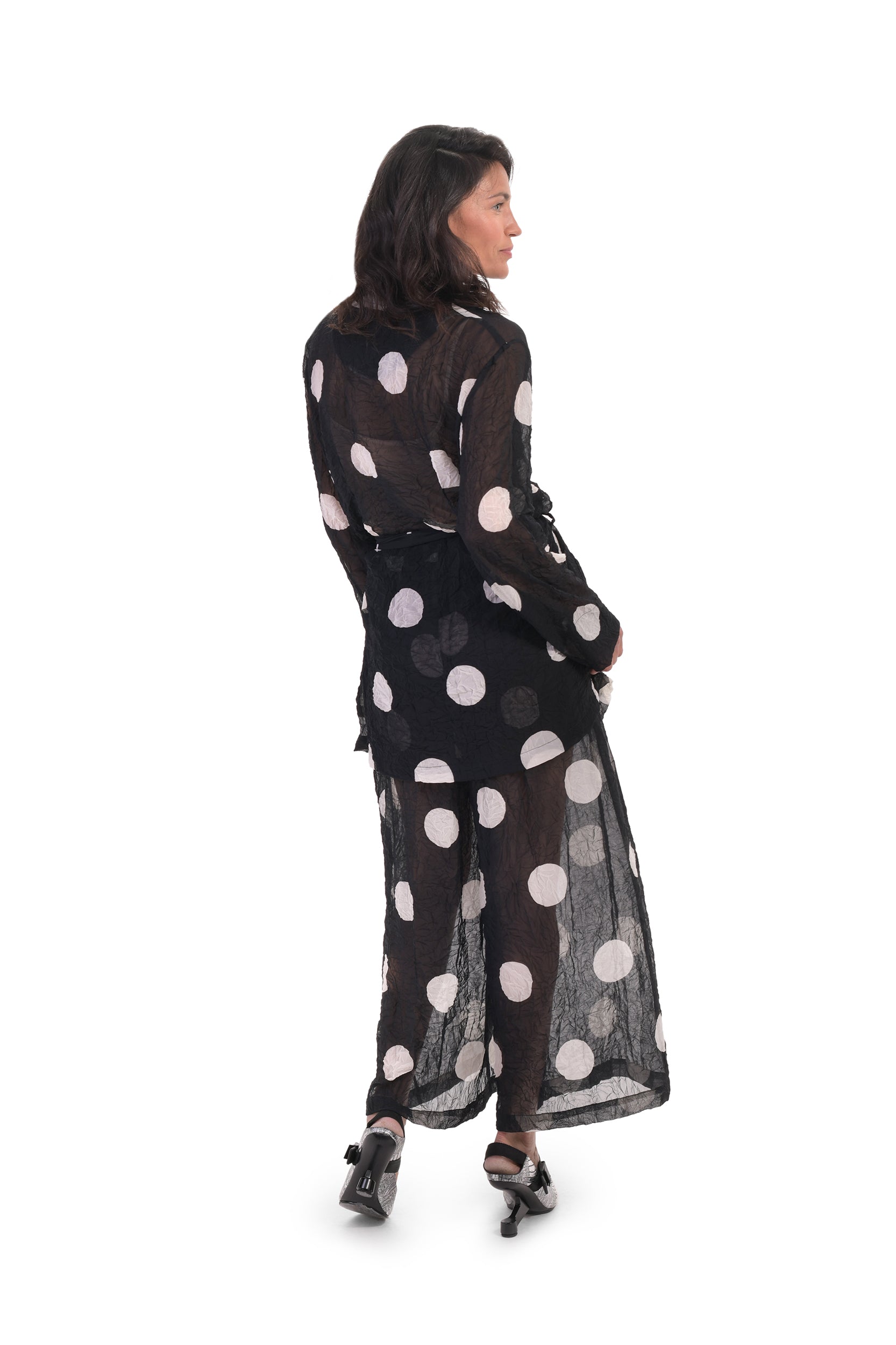 Back full body view of a woman wearing the alembika chiffon dot palazzo pant with a matching chiffon shirt. The pant and shirt is black with white dots. The fabric appears sheer. The pants are wide legged and end right above the ankles.