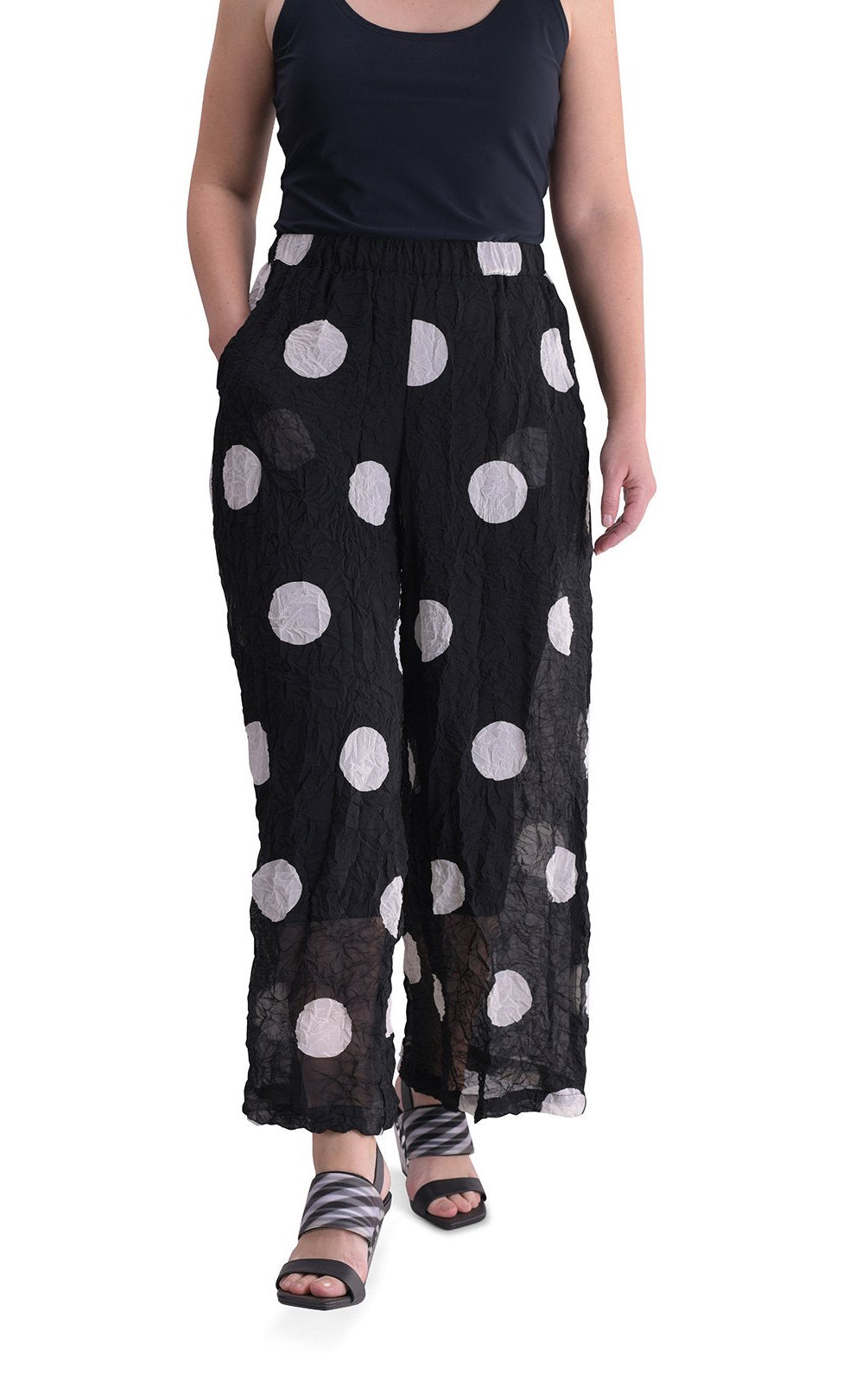 Front full body view of a woman wearing the alembika chiffon dot palazzo pant with a black tank. The pant is black with white dots. The fabric appears sheer. The pants are wide legged and end right above the ankles.