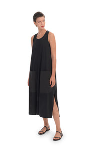 Front full body view of a woman wearing the alembika cotton tank dress. This black dress is sleeveless with two large patch pockets on the front and long side slits. This dress goes down to just below the model's knees.