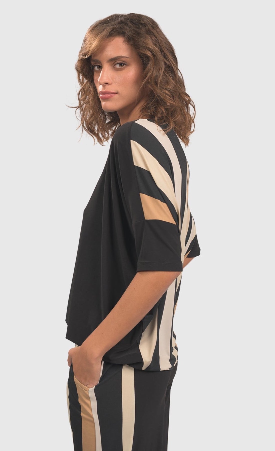 left side top half view of a woman wearing the alembika drapey dolman sunrise top. This top is black in the front and striped in the back with white and tan stripes. The top has elbow-length sleeves.