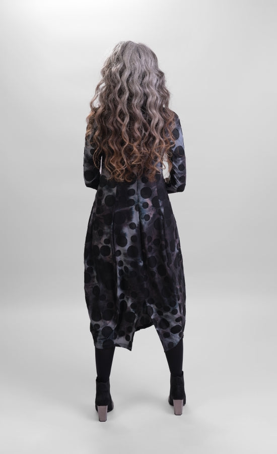 Back full body view of a woman wearing the alembika echo swing dress. This dress is grey, black, and blue tie dye with black dots all over it. It has a balloon skirt with a slit in the back and long sleeves.