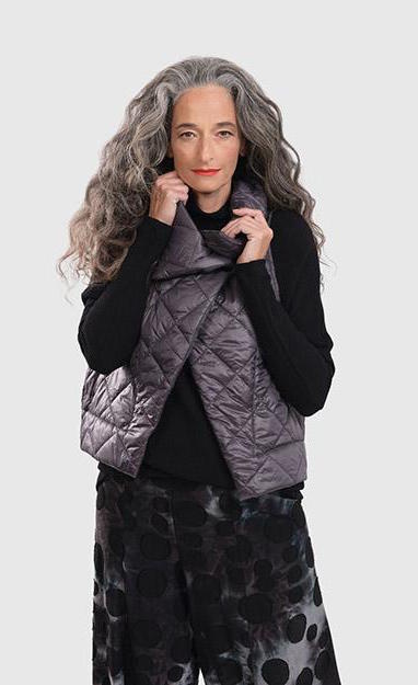 Front top half view of a woman wearing the alembika ether fina jacket vest in grey over a black long sleeve. This vest is a puffer vest. It features a large folded over collar and a single button closure towards the neck.