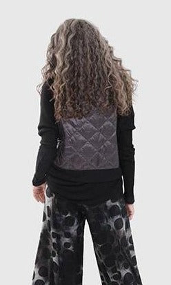 Back top half view of a woman wearing the alembika ether fina jacket vest in grey over a black long sleeve. This vest is a puffer vest. It features a large folded over collar and a single button closure towards the neck. The hem on the back is black and ribbed.
