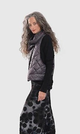 Left side top half view of a woman wearing the alembika ether fina jacket vest in grey over a black long sleeve. This vest is a puffer vest. It features a large folded over collar and a single button closure towards the neck. The hem on the back is black and ribbed.