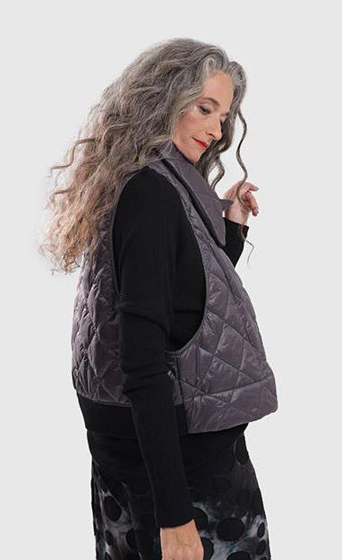 Right side top half view of a woman wearing the alembika ether fina jacket vest in grey over a black long sleeve. This vest is a puffer vest. It features a large folded over collar and a single button closure towards the neck. The hem on the back is black and ribbed. 