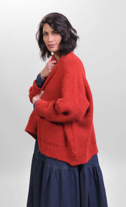 Left side top half view of a woman wearing the Alembika Signature Cardigan over a denim dress. This cardigan is red/flame colored. It has long sleeves, a 3-button front, and two front pockets.