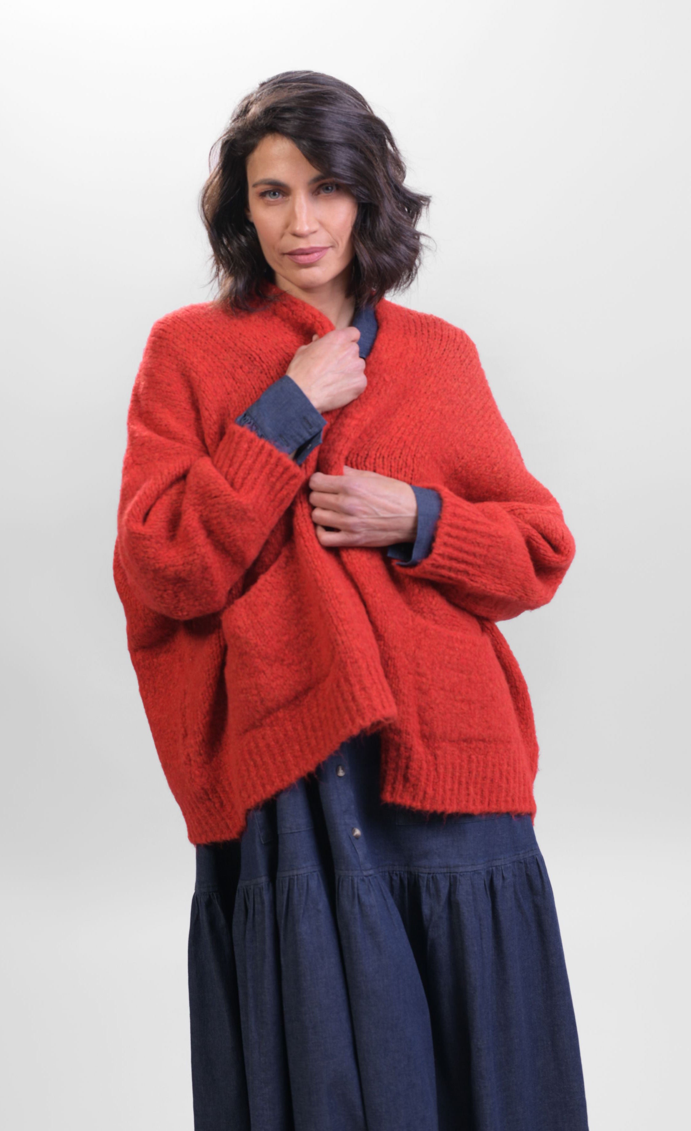 Front top half view of a woman wearing the Alembika Signature Cardigan. This cardigan is red/flame colored. It has long sleeves, a 3-button front, and two front pockets.